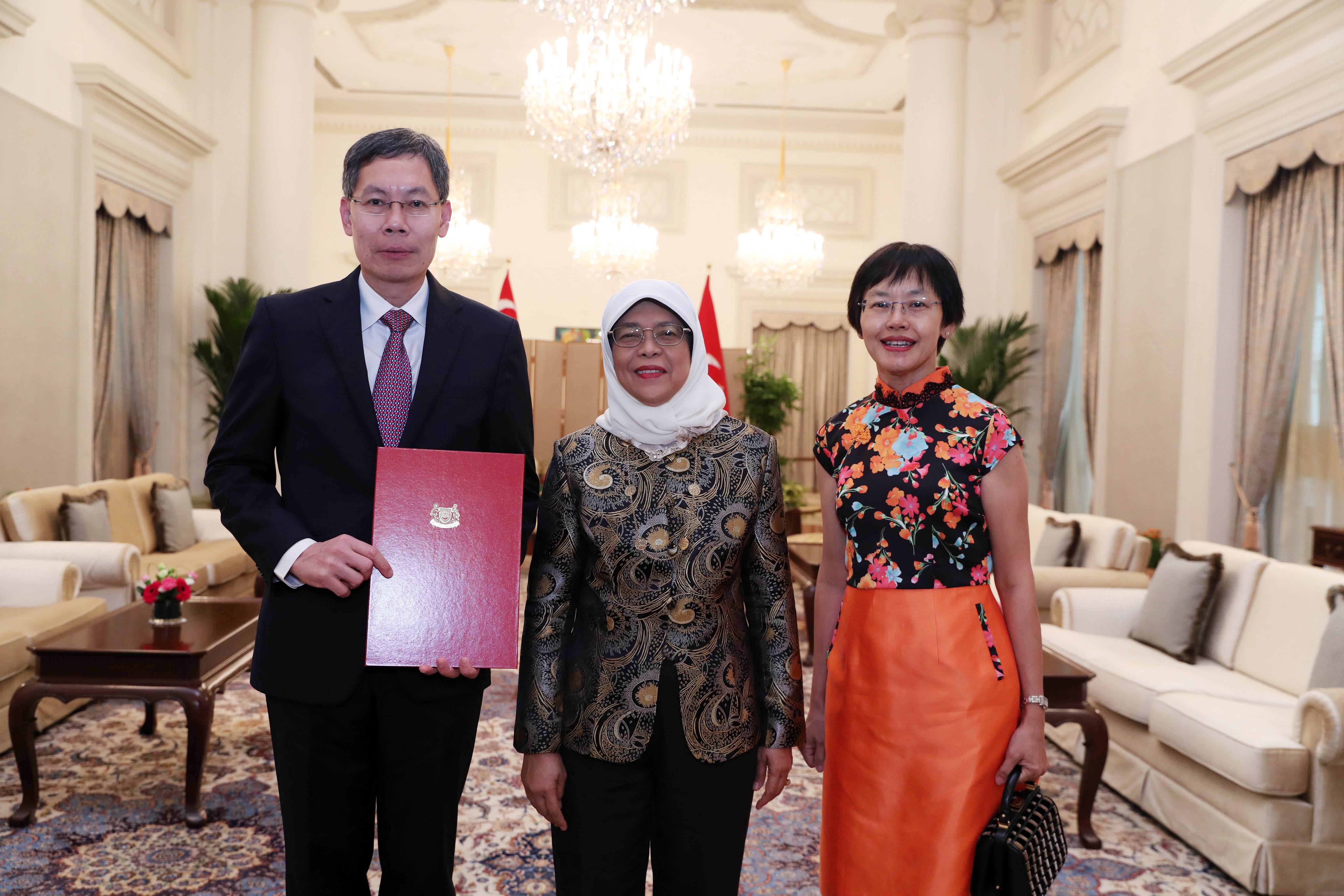 Three people stand indoors, one holding a red folder with Singapore's emblem.